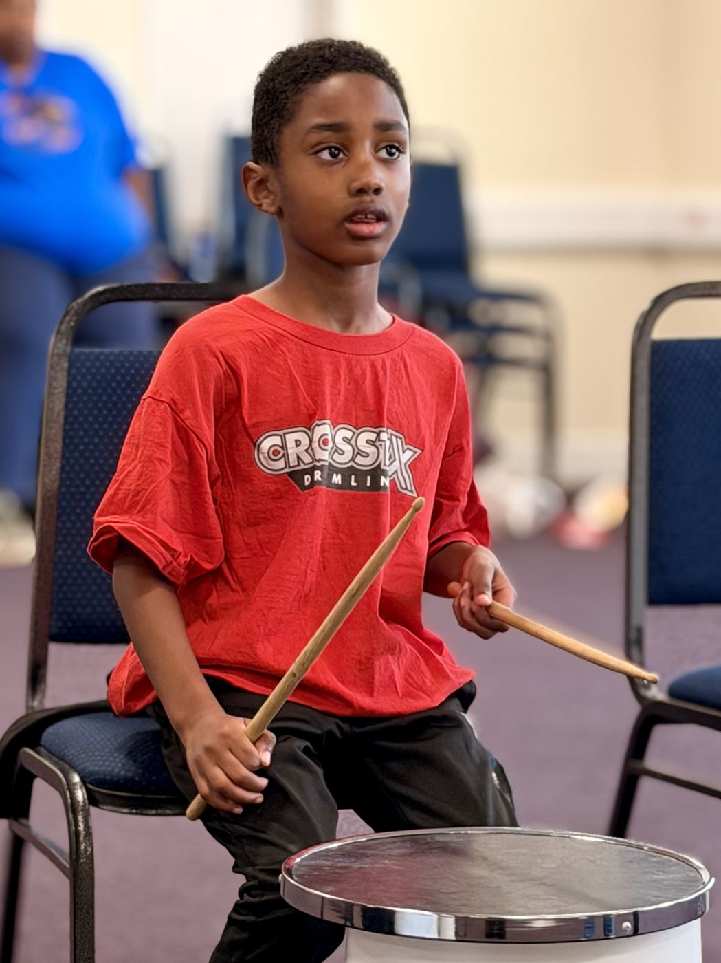 A young academy member sits at a practice drum, sticks ready, concentrating